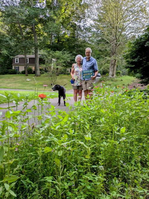A Connecticut couple and their dog, Bella, pose proudly with a Reduce Your Lawn Day sign in front of their pollinator-friendly strip. 